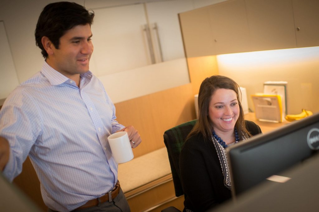 the woman is sitting at the computer smiling the man is standing is looking at the monitor and holding a white bucket