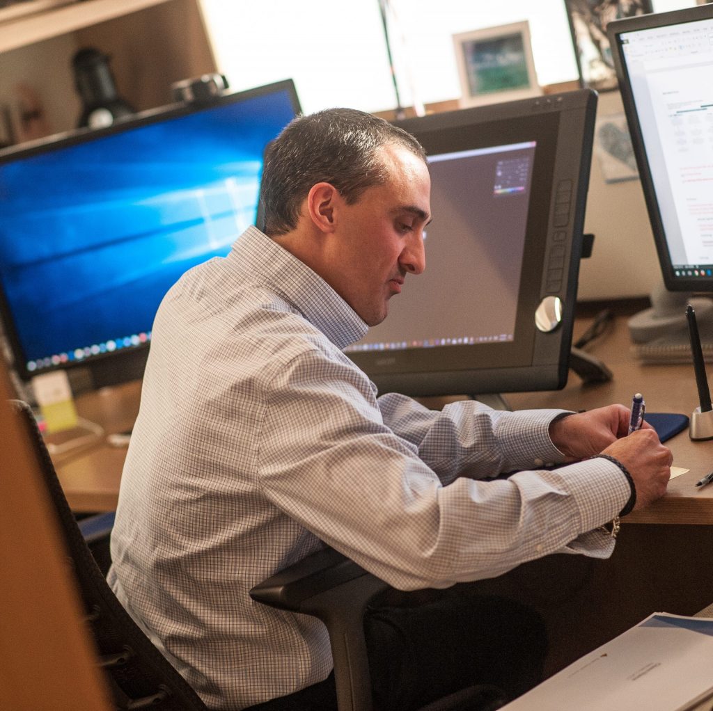 a man in a checked shirt writing on a piece of paper in front of the computer monitors