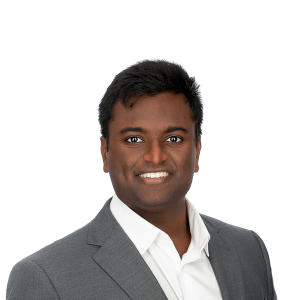 a dark skinned man with dark hair wearing a white shirt and gray jacket smiles at the camera