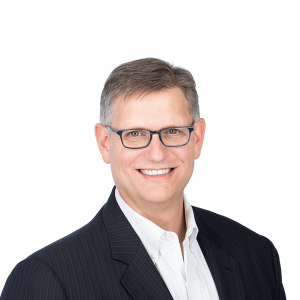 a man with gray hair wearing dark-rimmed glasses wearing a white shirt and a black striped suit smiles at the camera