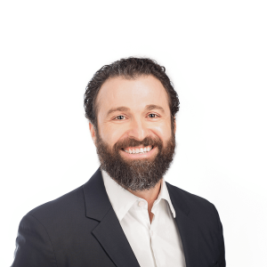 a man in dark hair with a beard, wearing a black jacket and white shirt smiles at the camera