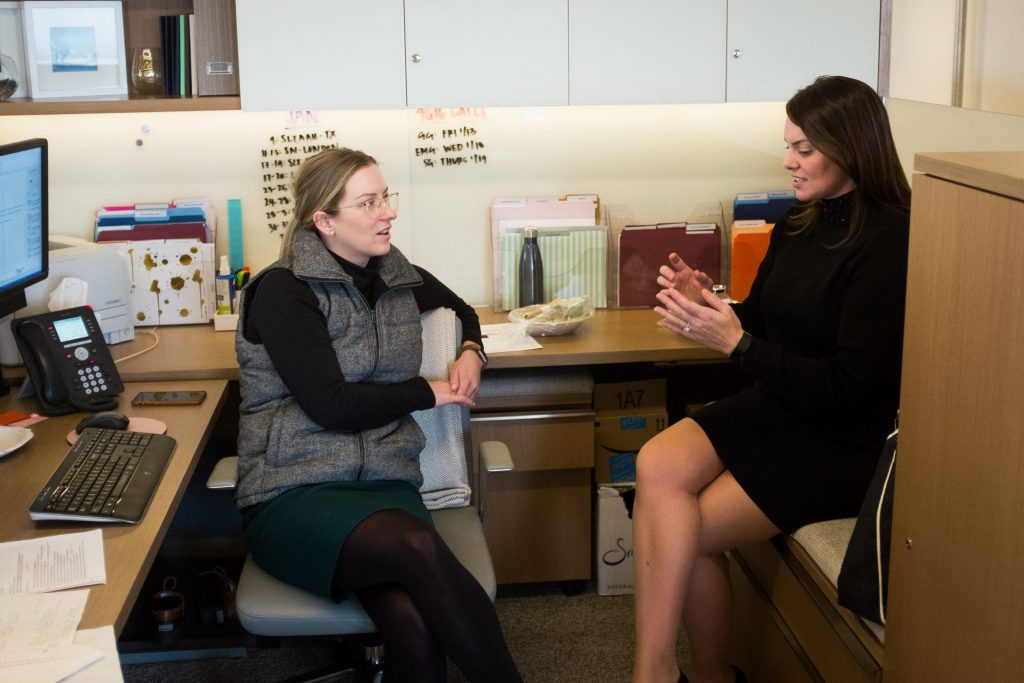 a woman in a vest and glasses is sitting in the office and talking to a woman in a black dress
