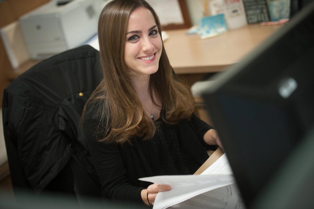 a woman with brown hair sitting at the computer smiles at the camera