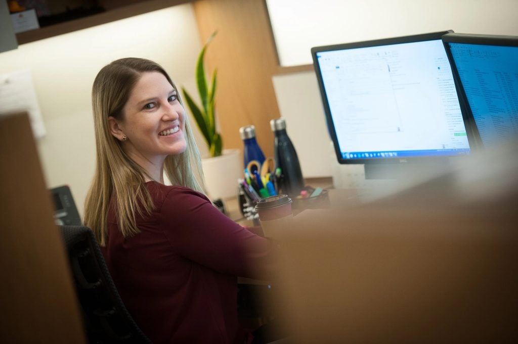 a woman with blonde hair wearing a brown sweater sitting in front of a computer smiles at the camera